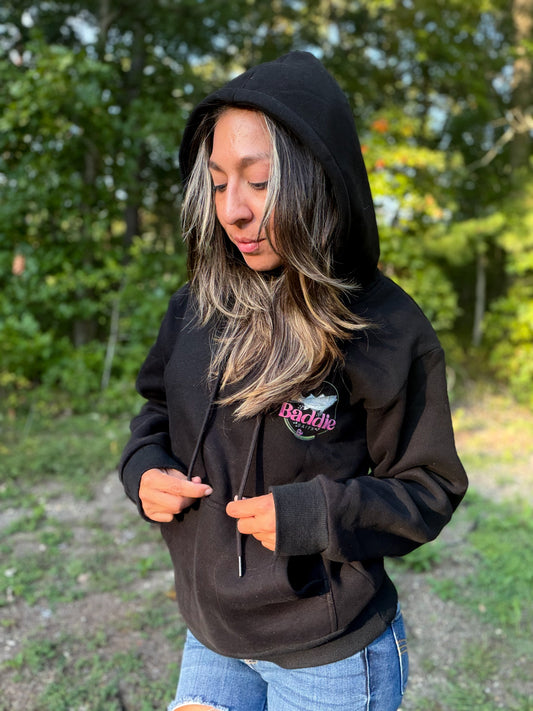 A woman wearing a black pullover hoodie with a pink and white 'Bad Blood' graphic on the left chest, paired with blue jeans, standing outdoors in front of a blurred green forest background.